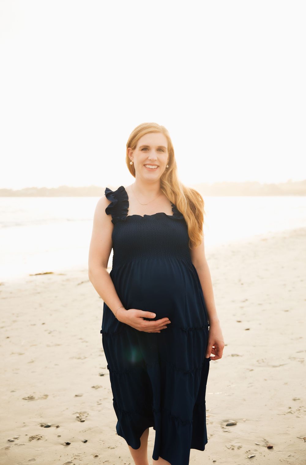 Smiling pregnant woman in navy dress cradling baby bump on Second Beach Middletown, RI at golden hour.