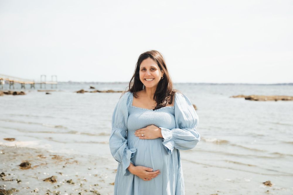 Pregnant woman in a light blue dress cradling her baby bump on a rocky beach by the ocean. maternity photoshoot, Beavertail Jamestown RI