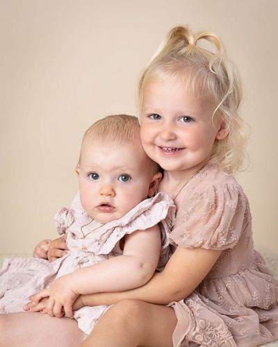 Two young children in soft pink dresses pose together for a portrait against a neutral beige background.