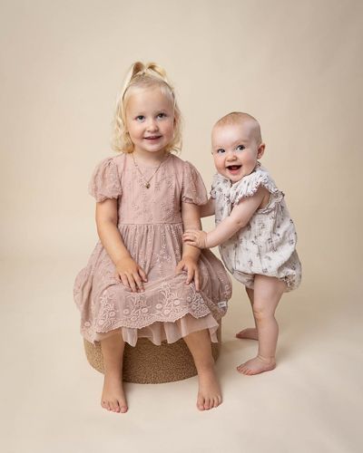 Two children in soft pink and floral outfits sit together on a neutral beige backdrop for a portrait session.