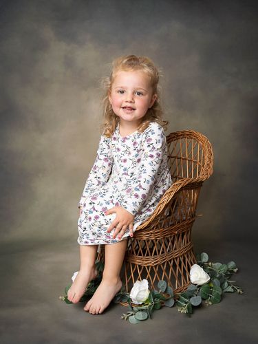 A child in a floral dress poses on a wicker chair with white roses and eucalyptus decorations in a studio setting.