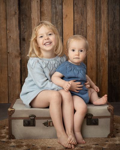 Siblings sitting together on wooden bench against rustic barn wall backdrop.
