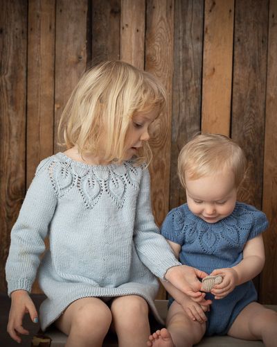 Two children in matching knit sweaters sit together against a rustic wooden wall background.