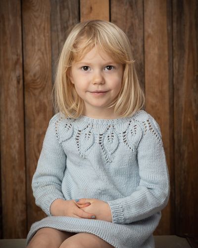 A child in a light blue knitted sweater with a delicate leaf pattern sits against a rustic wooden wall background.