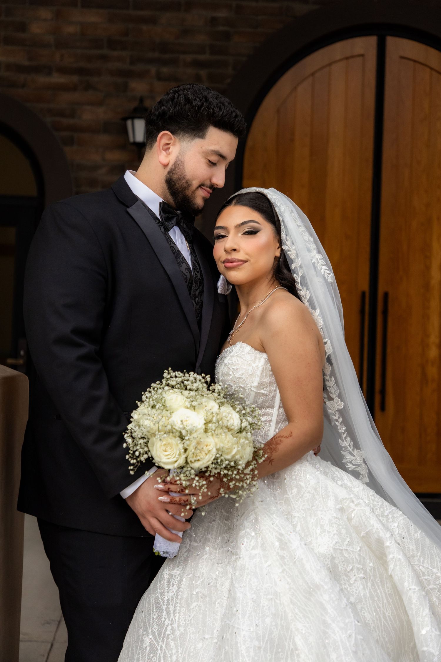 Bride in a strapless lace ball gown and groom in black tuxedo pose together in front of wooden doors.