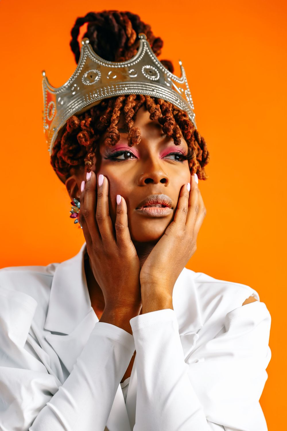 Person wearing ornate crown and white outfit poses dramatically against vibrant orange background.