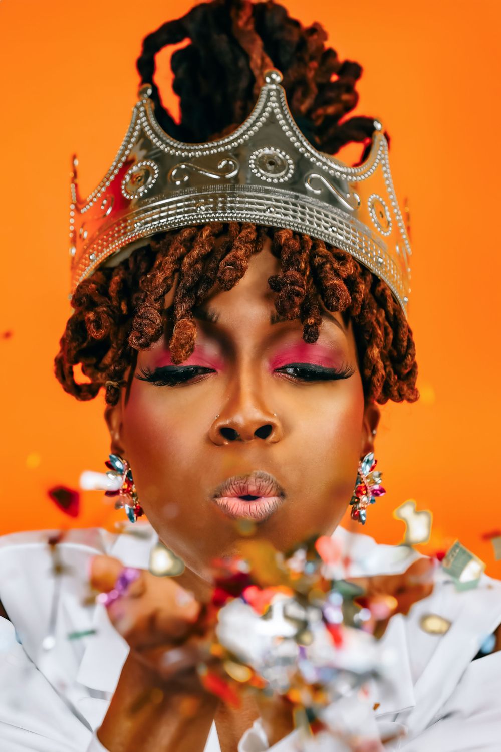 Person in white outfit and crown blowing silver confetti against bright orange backdrop.