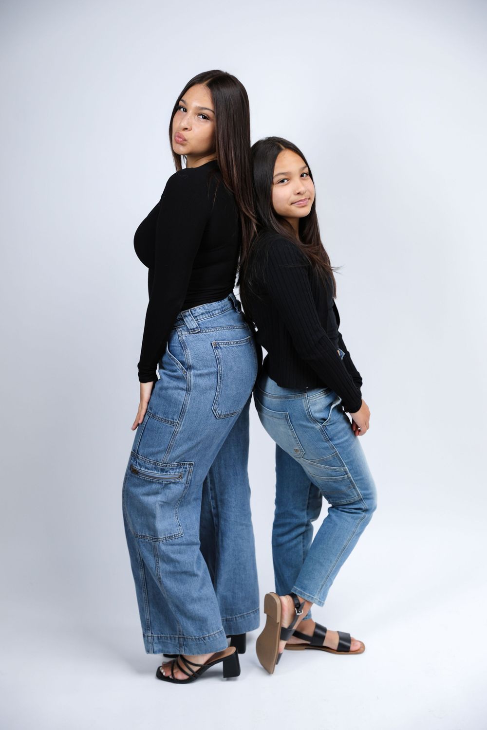 Two people wearing black tops and blue jeans pose casually on the floor in a studio setting.