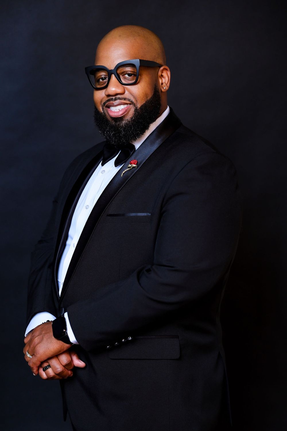 Professional headshots of person in formal black tuxedo and glasses against dark background.