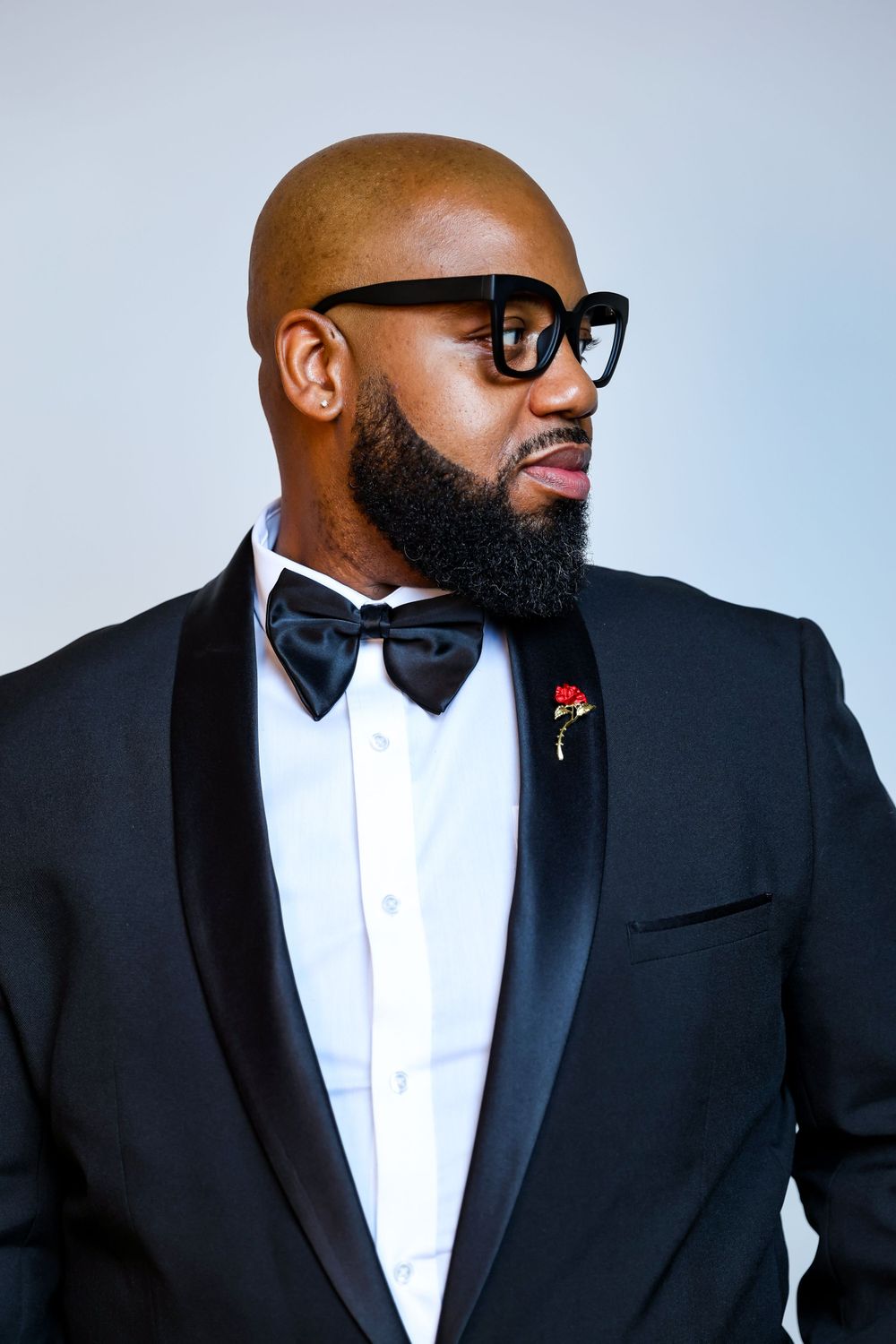 A dapper gentleman in a black tuxedo, bow tie and glasses poses for a formal portrait.