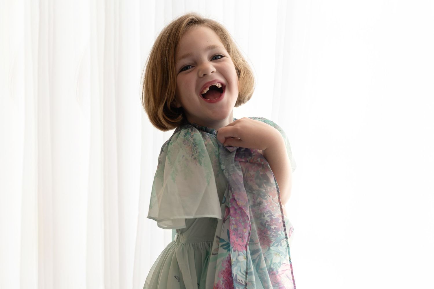 Young child wearing a flowing mint green and floral dress laughs joyfully against a bright background.