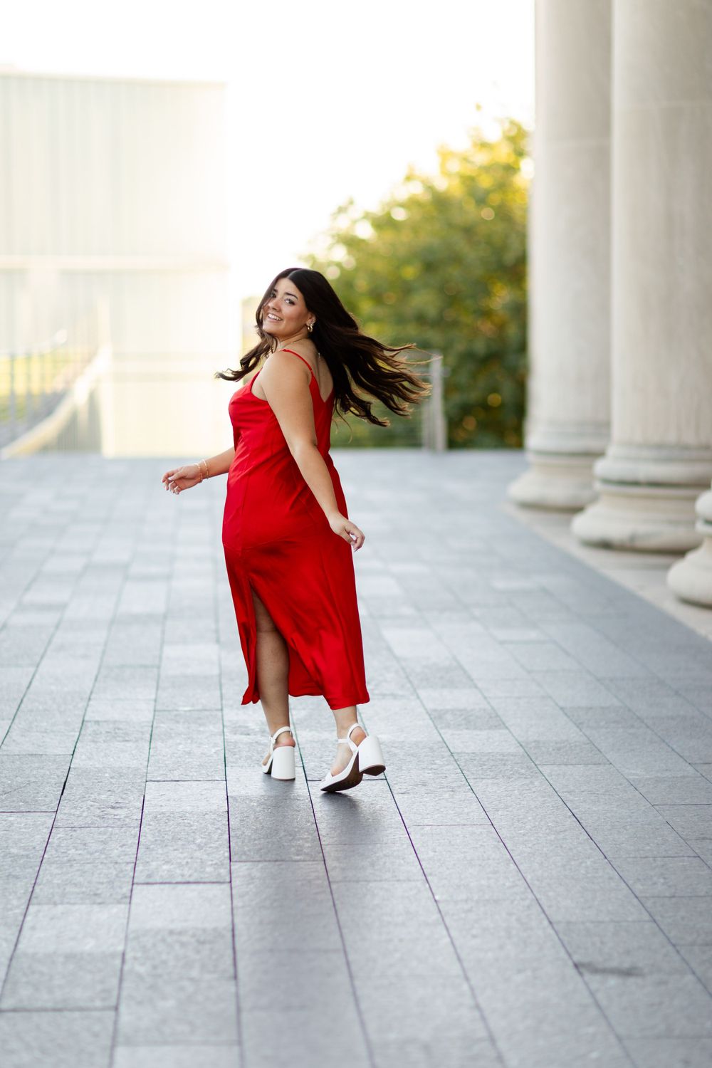 A woman in a red dress twirls playfully between white columns during a warm sunset photoshoot.