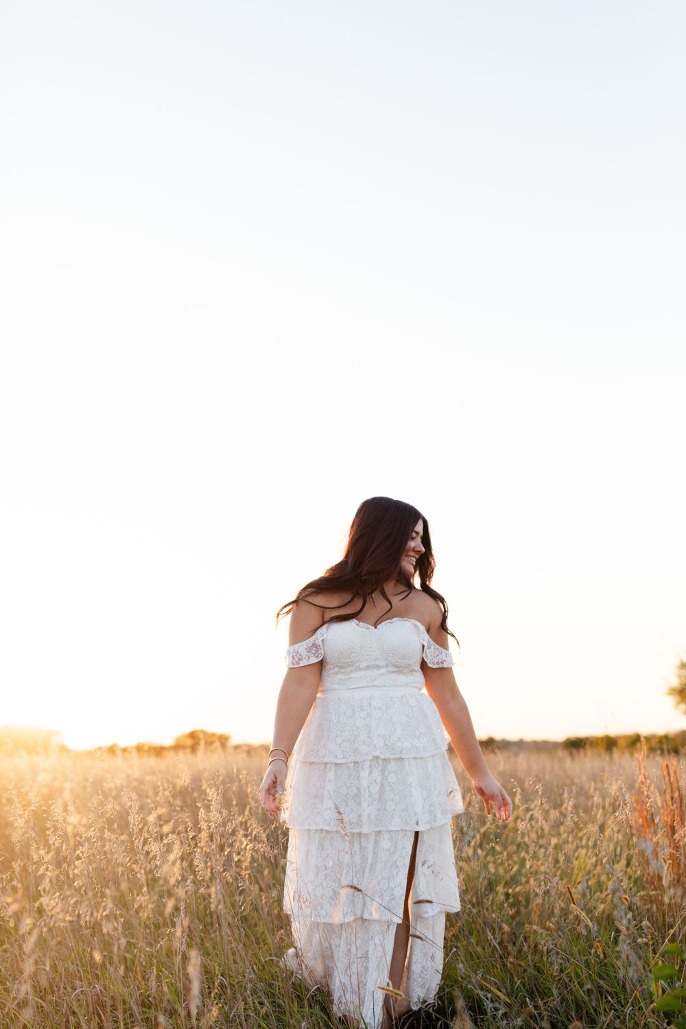 Silhouette in a tiered white dress stands gracefully in a sun-drenched meadow.