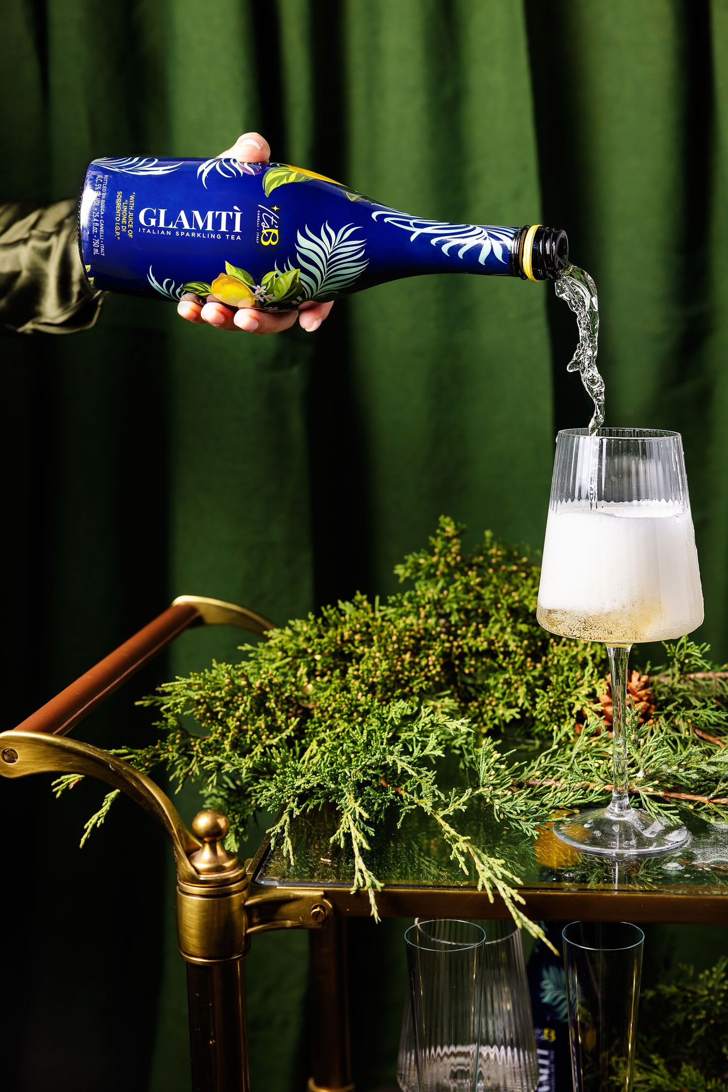 Hand pouring Glamti sparkling wine into a glass on a gold bar cart with greenery.