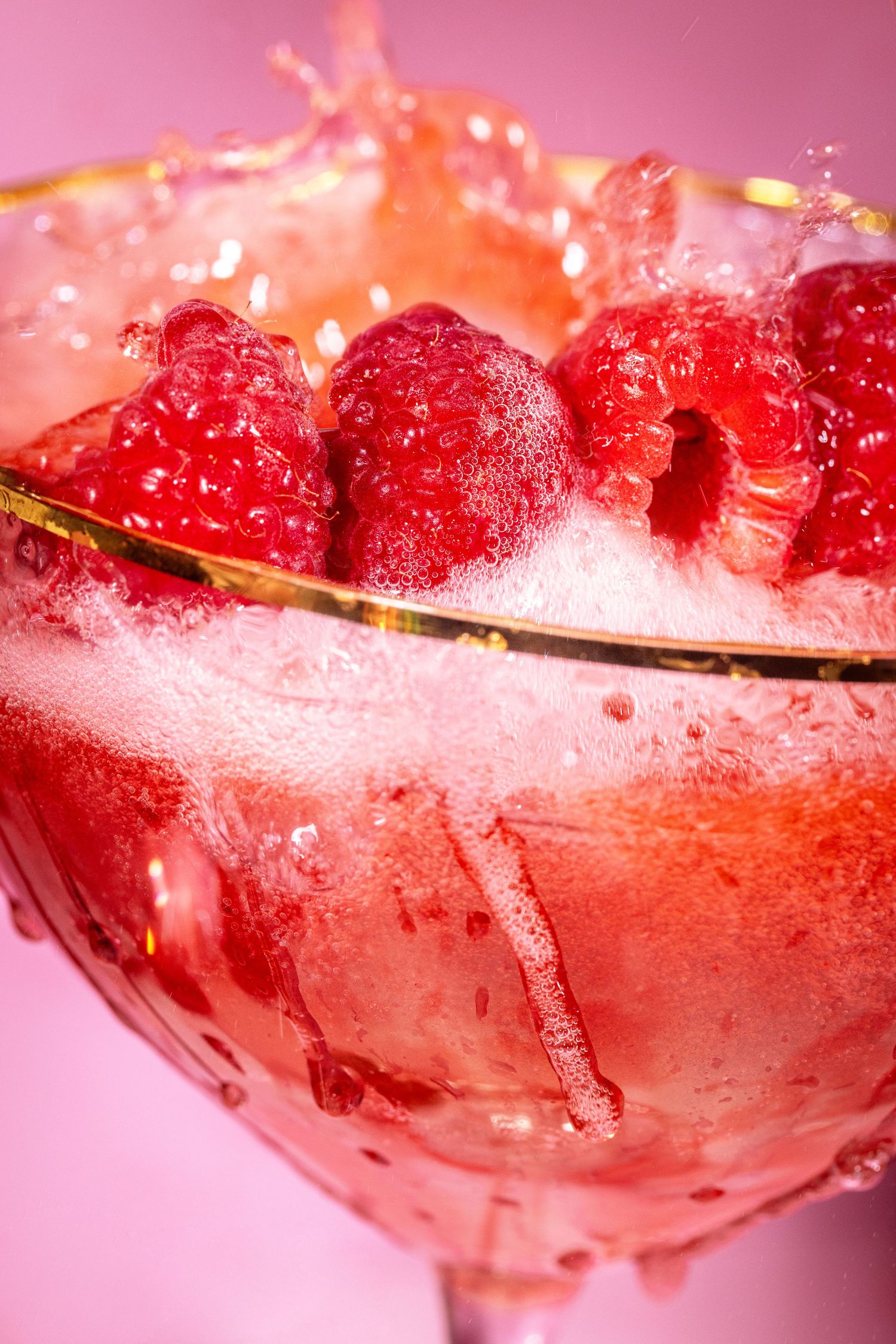 Fresh raspberries splashing in a gold-rimmed champagne coupe glass with pink sparkling liquid.