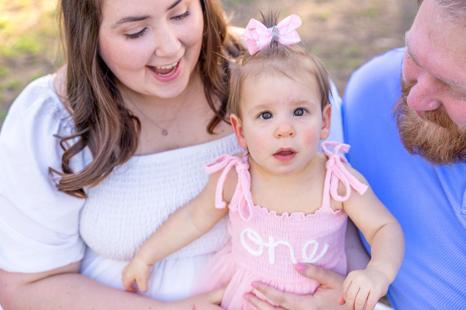Baby in pink dress with matching hair bow poses with her parents during her Raleigh outdoor family photoshoot.