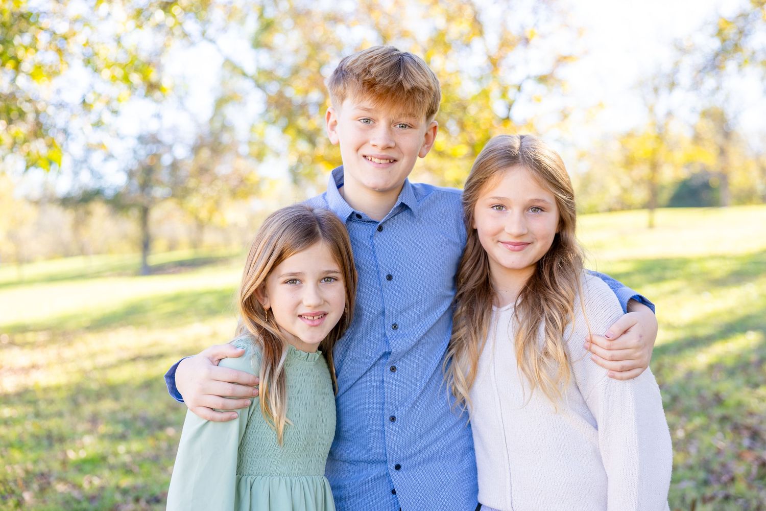 Three siblings pose together outdoors with autumn foliage in the background on a sunny day at Historic Oak View Park in Raleigh.