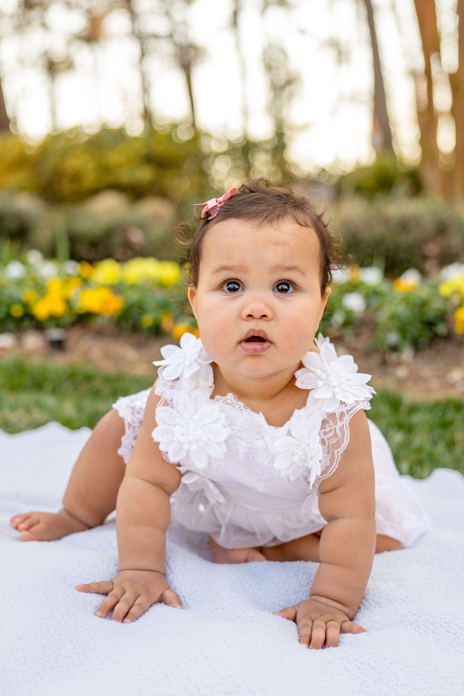 Baby in white ruffled outfit laying on white blanket with colorful garden flowers in background at WRAL Azalea Gardens in Raleigh.