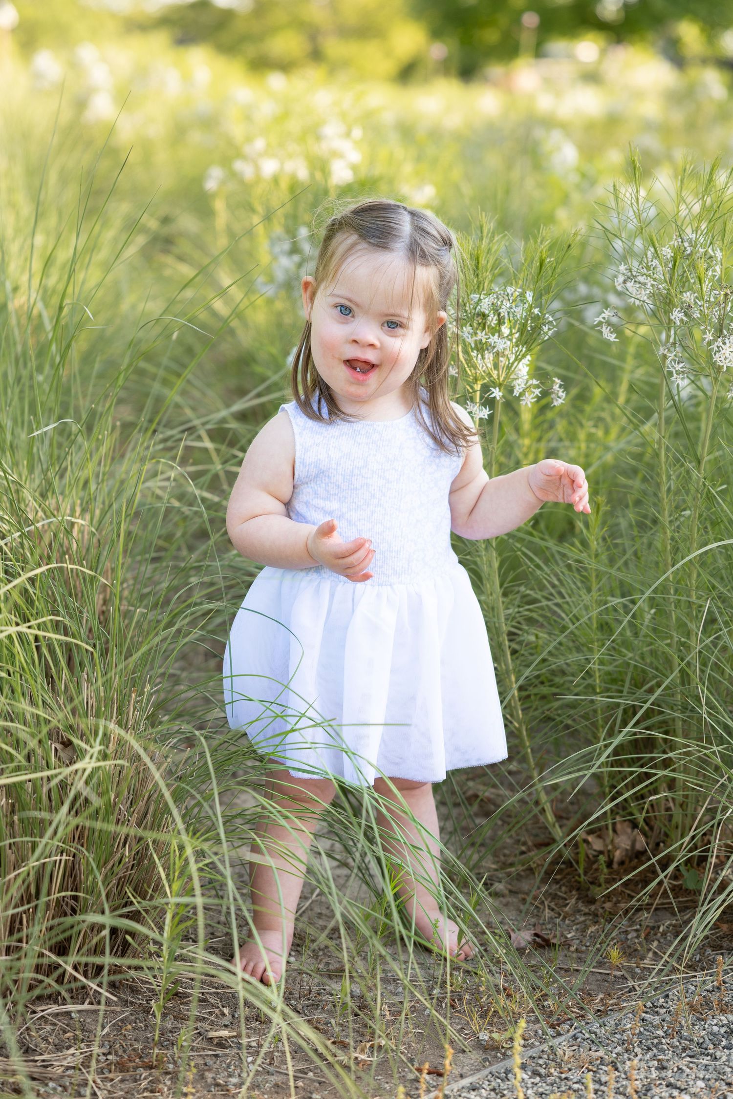 A 2 year old girl in a blue dress stands in a field of long grass smiling.