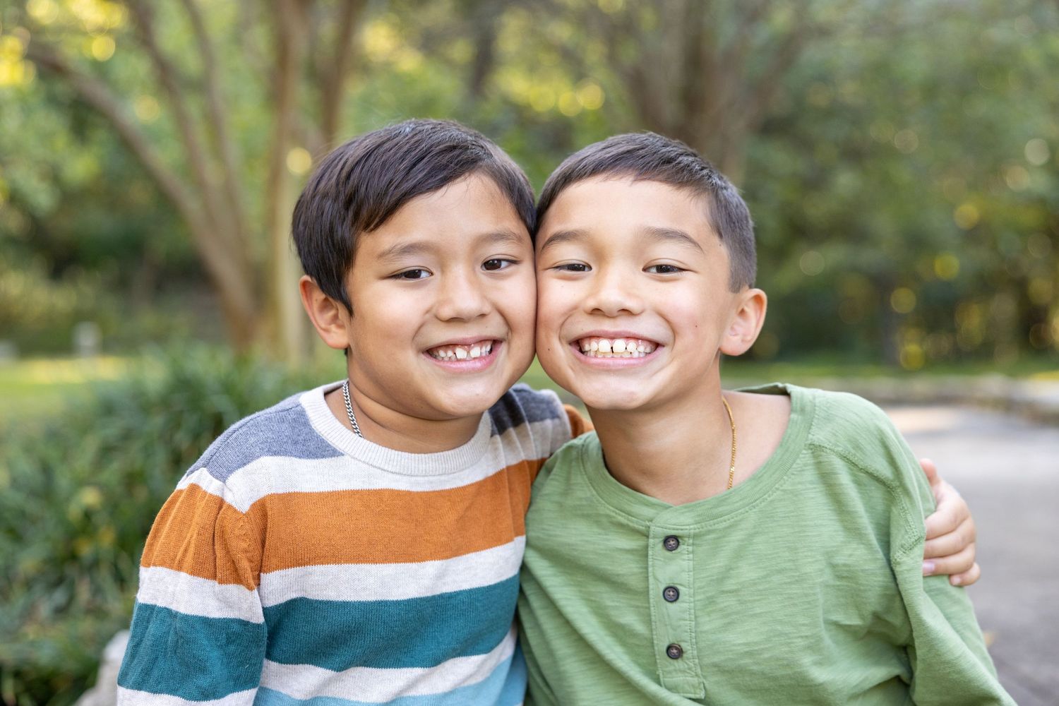 Two young boys in casual sweaters smile warmly together while embracing outdoors in a natural setting.