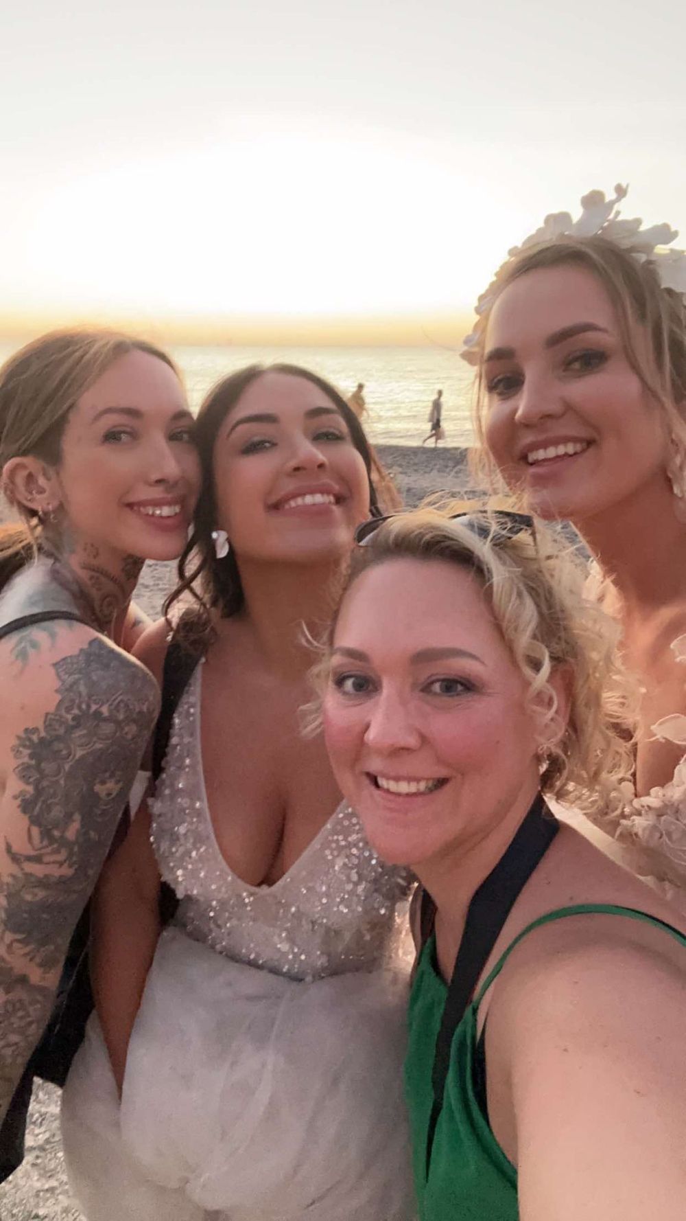 Happy group takes a sunset selfie at the beach in Greece with someone walking along the shoreline in the background.