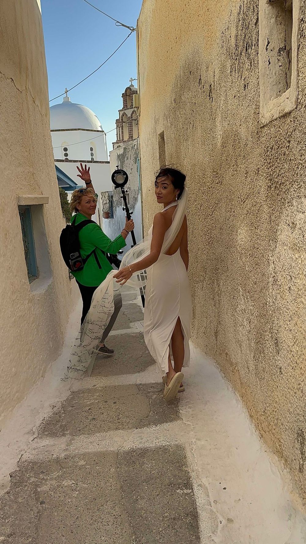 Two people pose playfully in a narrow whitewashed alley with a church dome and tower visible in the background in Greece.