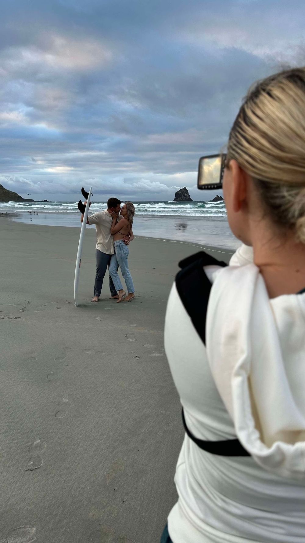 A photographer captures a couple on a cloudy beach with dramatic rock formations in the background.