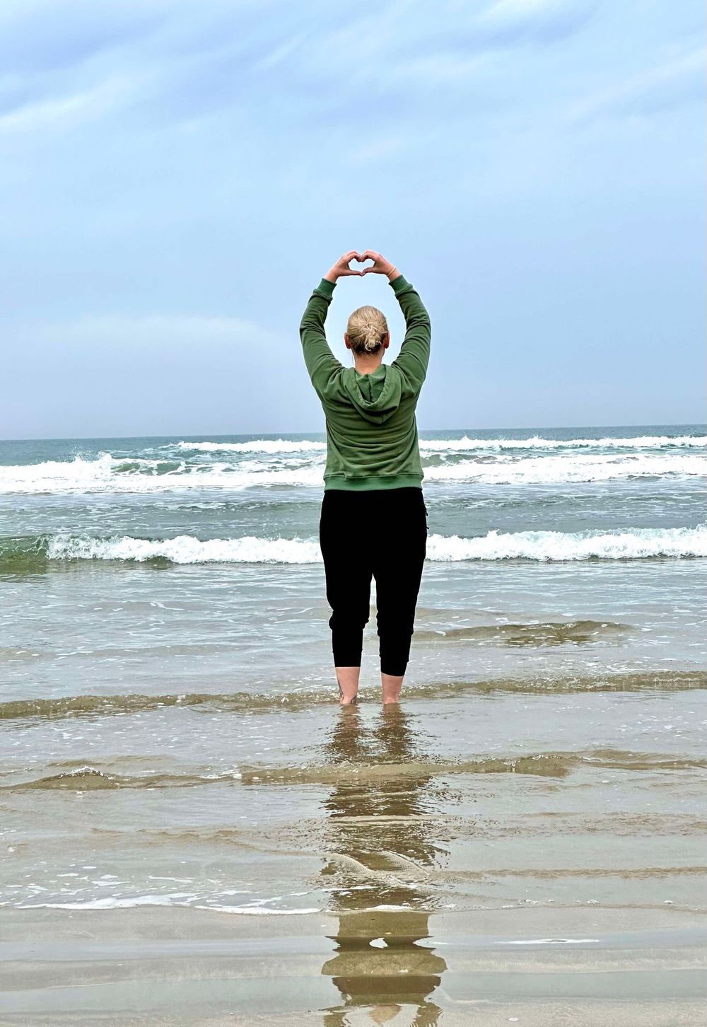 Person in green sweater making heart shape with arms while standing at the edge of ocean waves on beach.