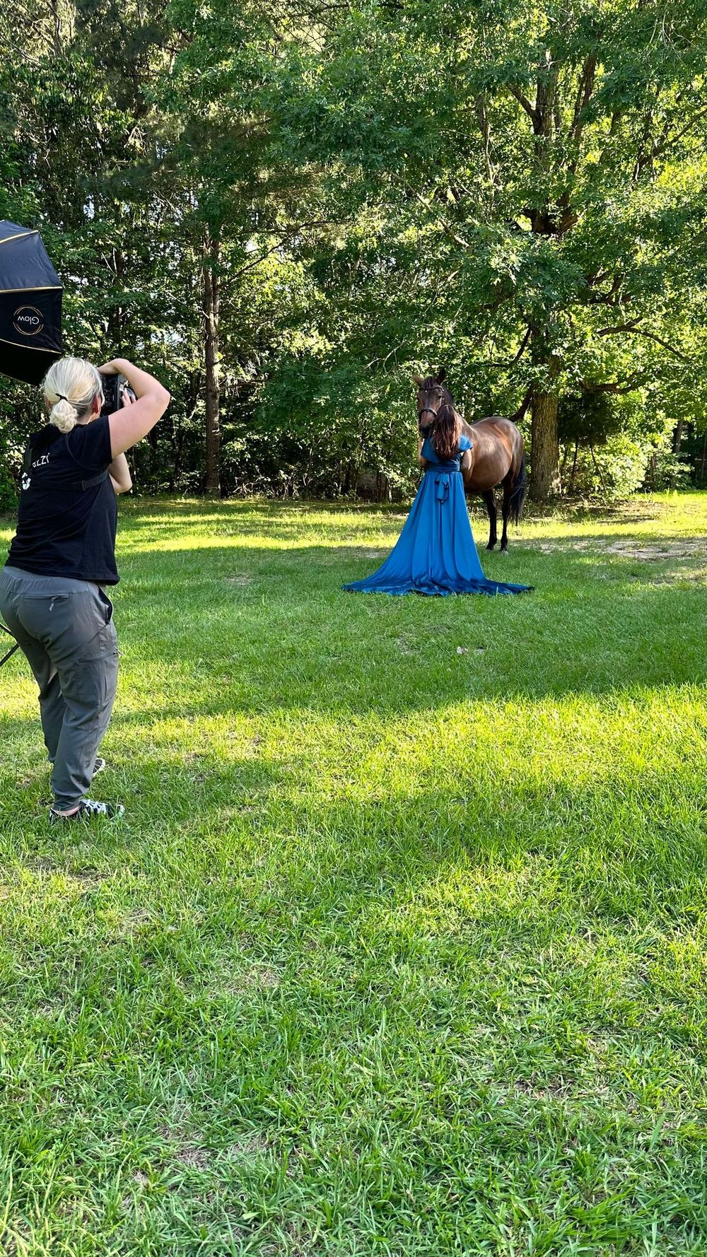 A photographer captures a photo shoot of someone in a flowing blue dress posing with a horse in a sunlit garden setting.