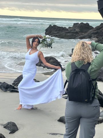 A photographer captures a photoshoot of someone in a flowing white dress on a rocky beach at sunset in New Zealand.