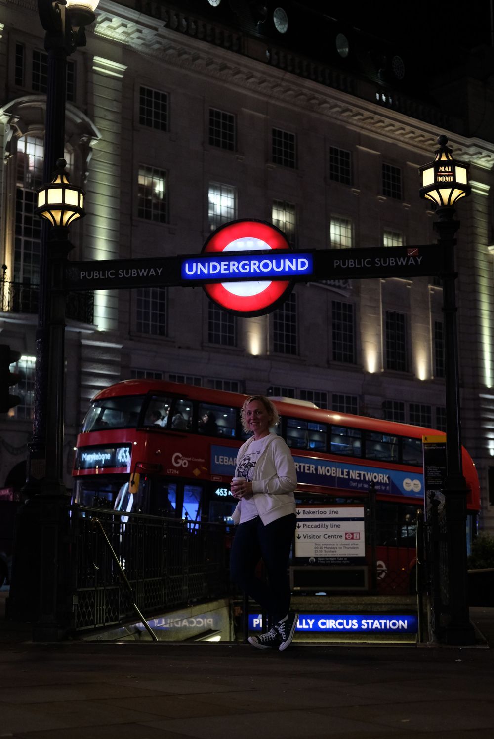 A London Underground station entrance at night with an illuminated sign and red double-decker bus outside Piccadilly Circus.