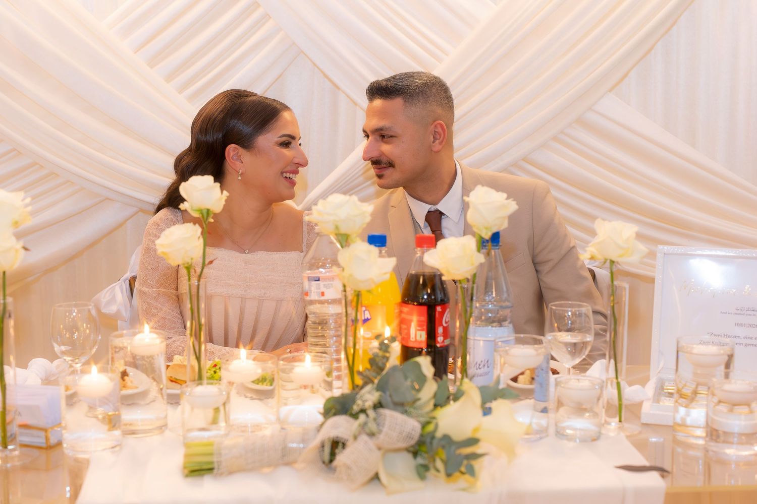 Happy couple at wedding reception table decorated with white roses, candles, and elegant floral arrangements.