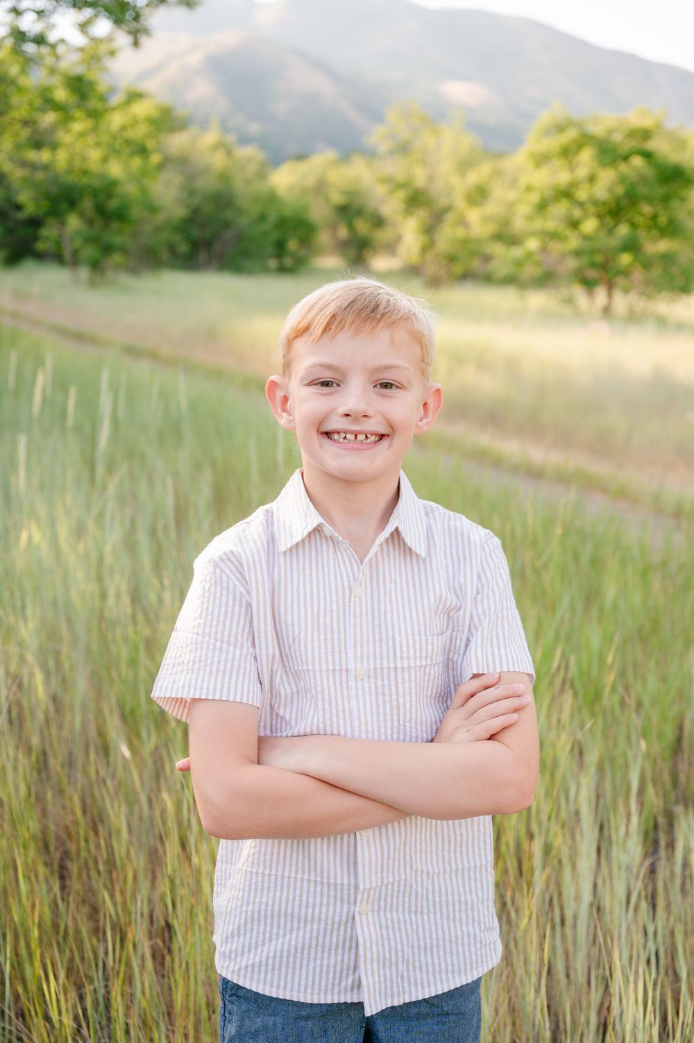 Smiling red-haired boy with crossed arms stands in a green field with mountains in the background.