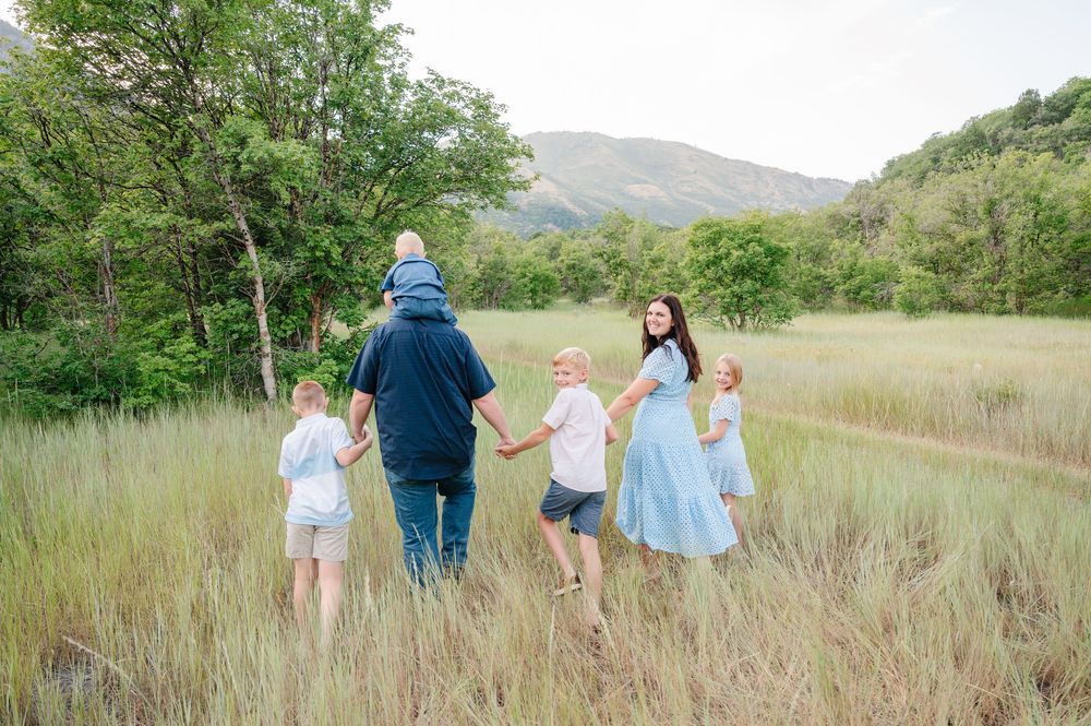 Family of six walking through a lush green meadow with mountains in the background on a sunny day.