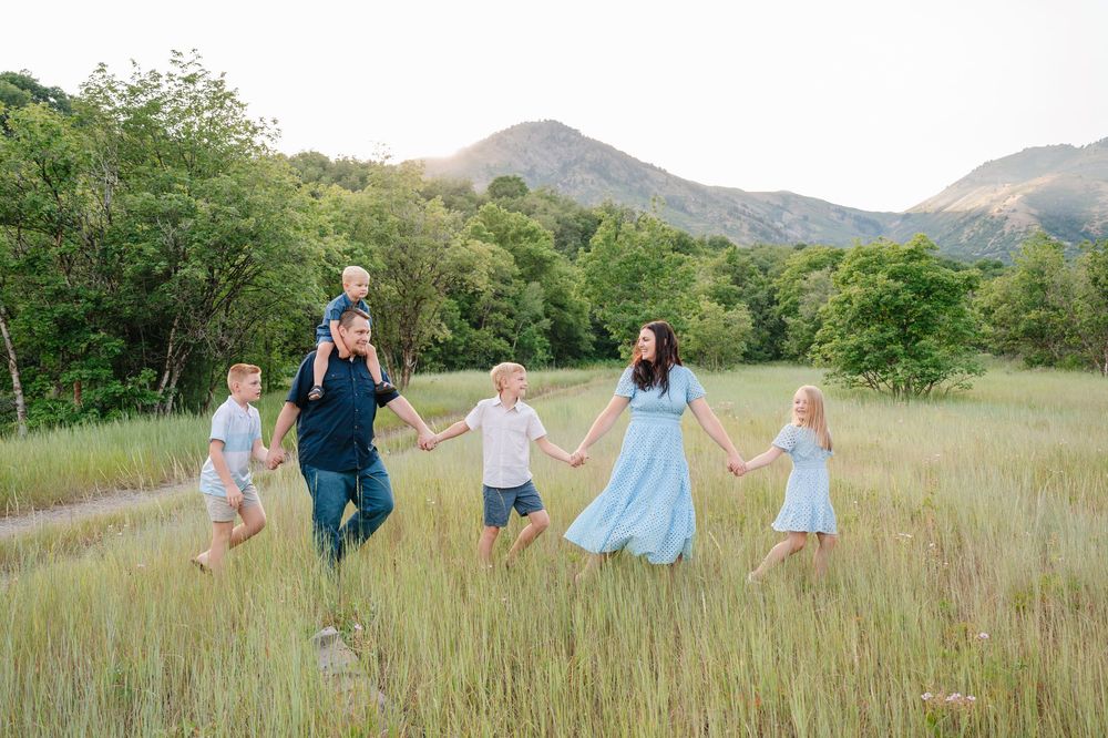Family of six walking hand-in-hand through a lush green meadow with mountains in the background.
