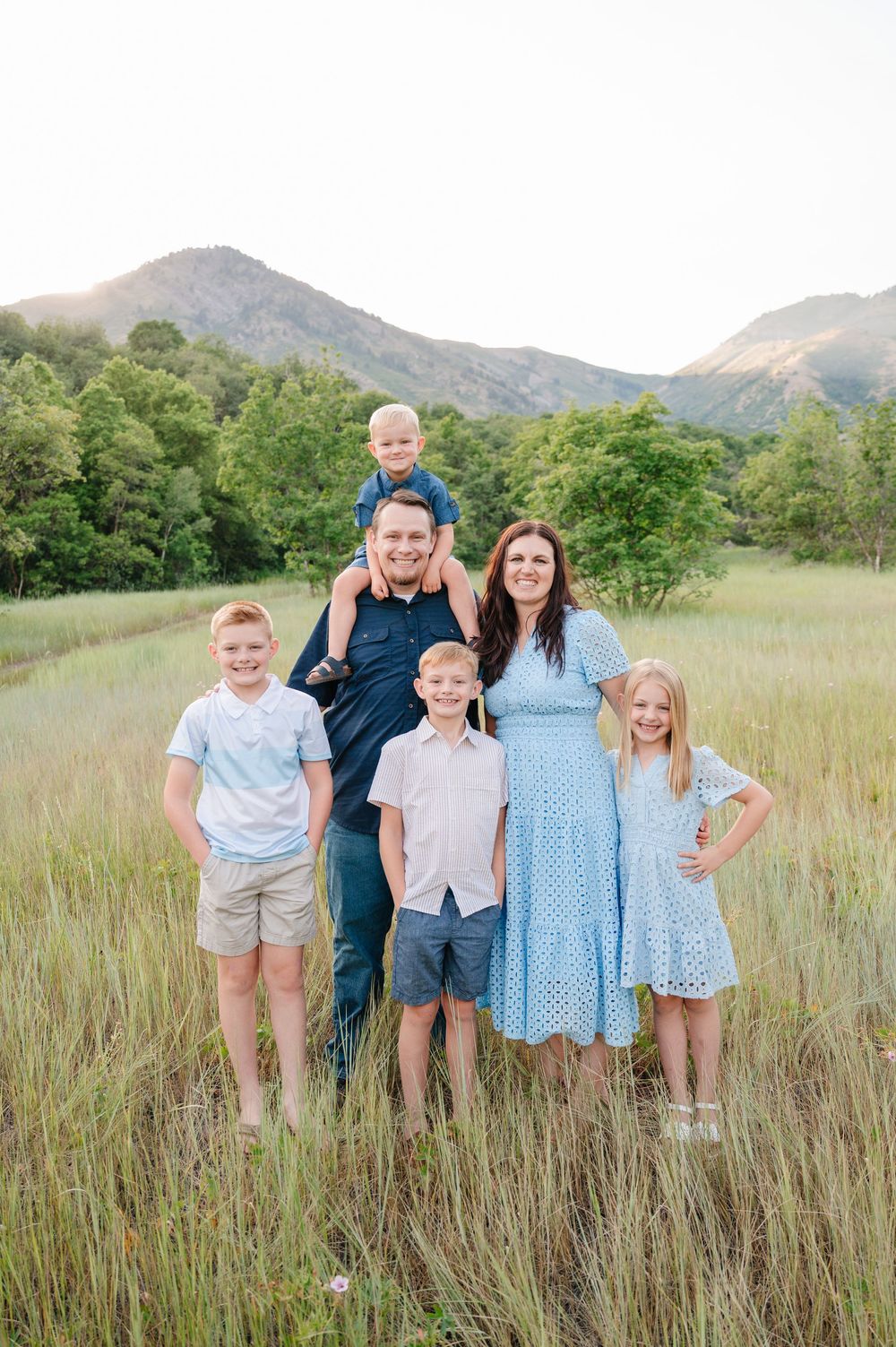 Family of six poses in a lush green meadow with mountain backdrop, parents and four children in coordinating blue outfits.