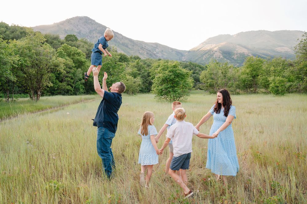 Family of five enjoying a sunny day in a mountain meadow, with dad tossing toddler in the air.