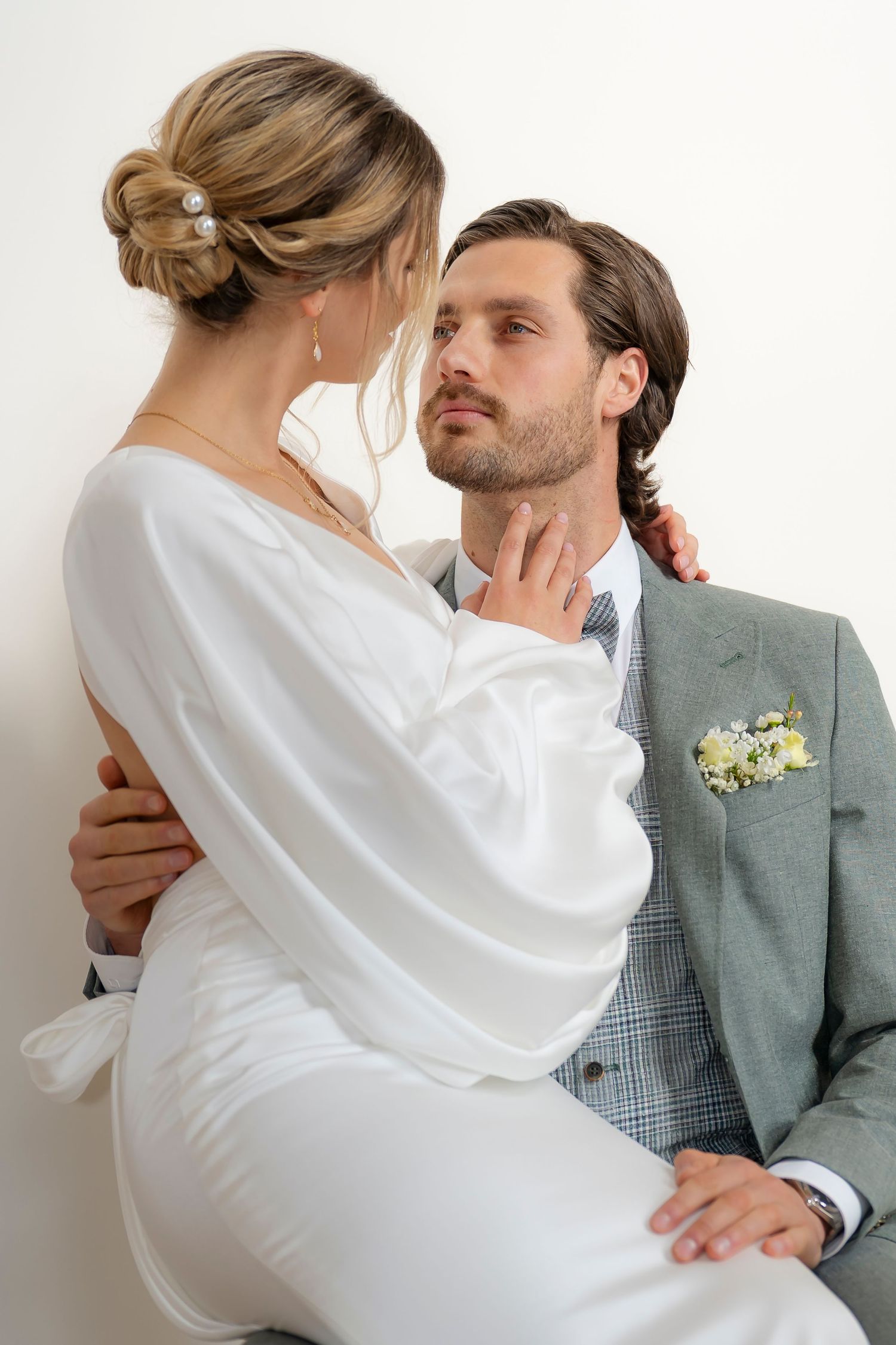 Bride in white satin gown sits on grooms lap as they share an intimate moment against a white wall.