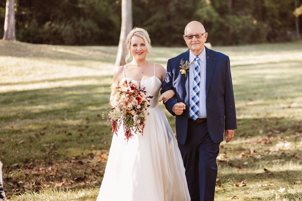 Bride in white gown holding red bouquet walks with elderly man in navy suit on green lawn.