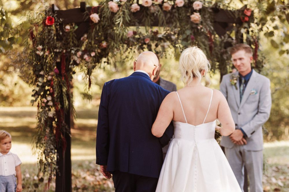 Bride walks down outdoor aisle with escort toward groom at floral arch ceremony.