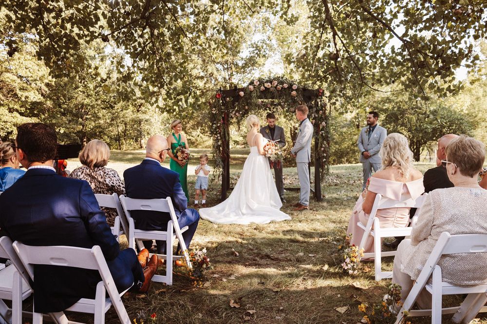 Outdoor wedding ceremony under floral arch with guests seated on white chairs among lush green trees.