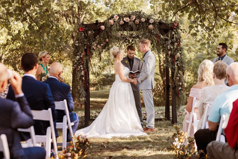 Couple exchanges vows at outdoor wedding ceremony under floral arch surrounded by seated guests in wooded setting.