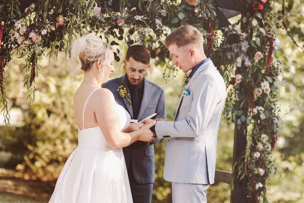 Groom reads vows to bride at floral wedding arch with officiant outdoors.