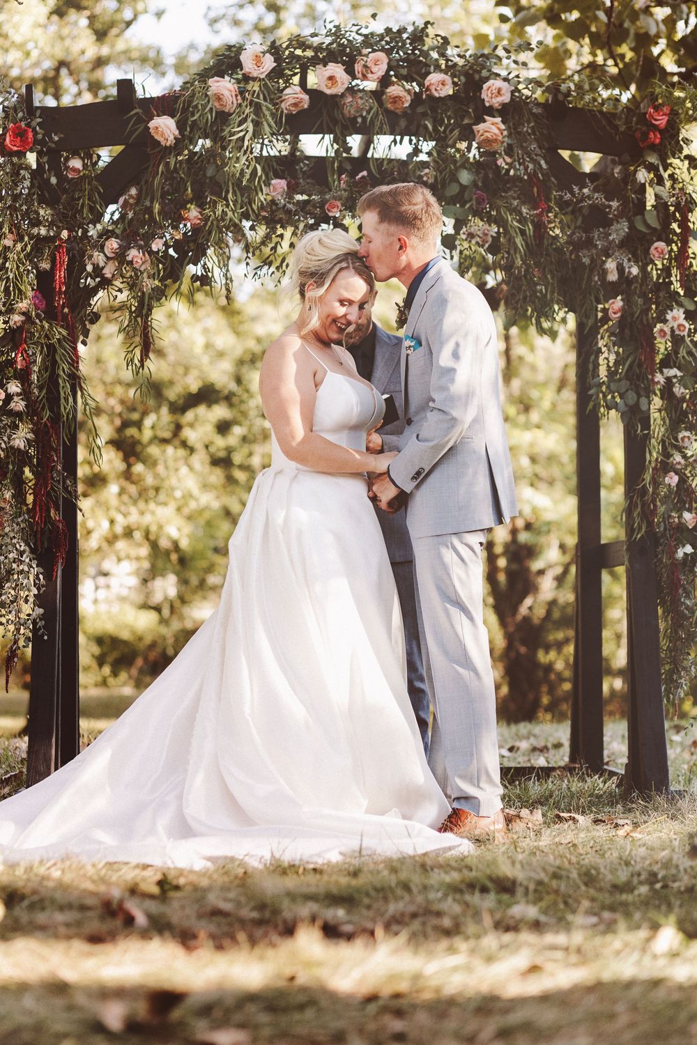 Bride and groom share a tender kiss under a floral arch adorned with pink roses at an outdoor wedding ceremony.