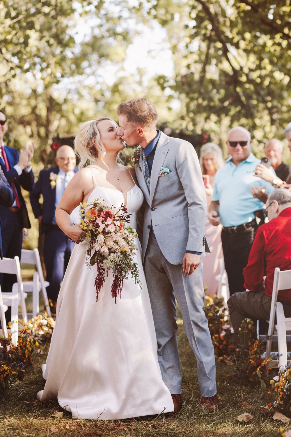 Bride and groom share a kiss at outdoor fall wedding ceremony while guests celebrate along the aisle.