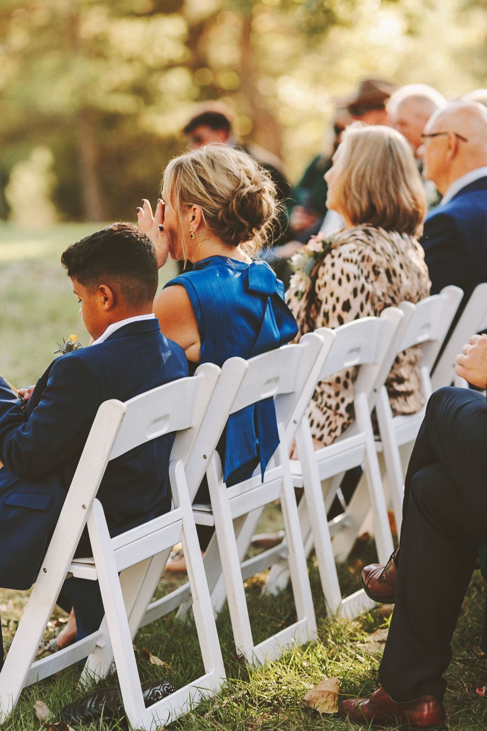 Guests seated in white folding chairs at an outdoor wedding ceremony, woman in blue dress wiping tears.