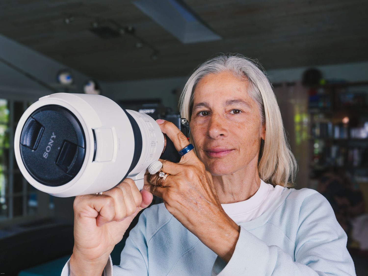 Older woman with gray hair holding a large white Sony telephoto camera lens up to her eye indoors.