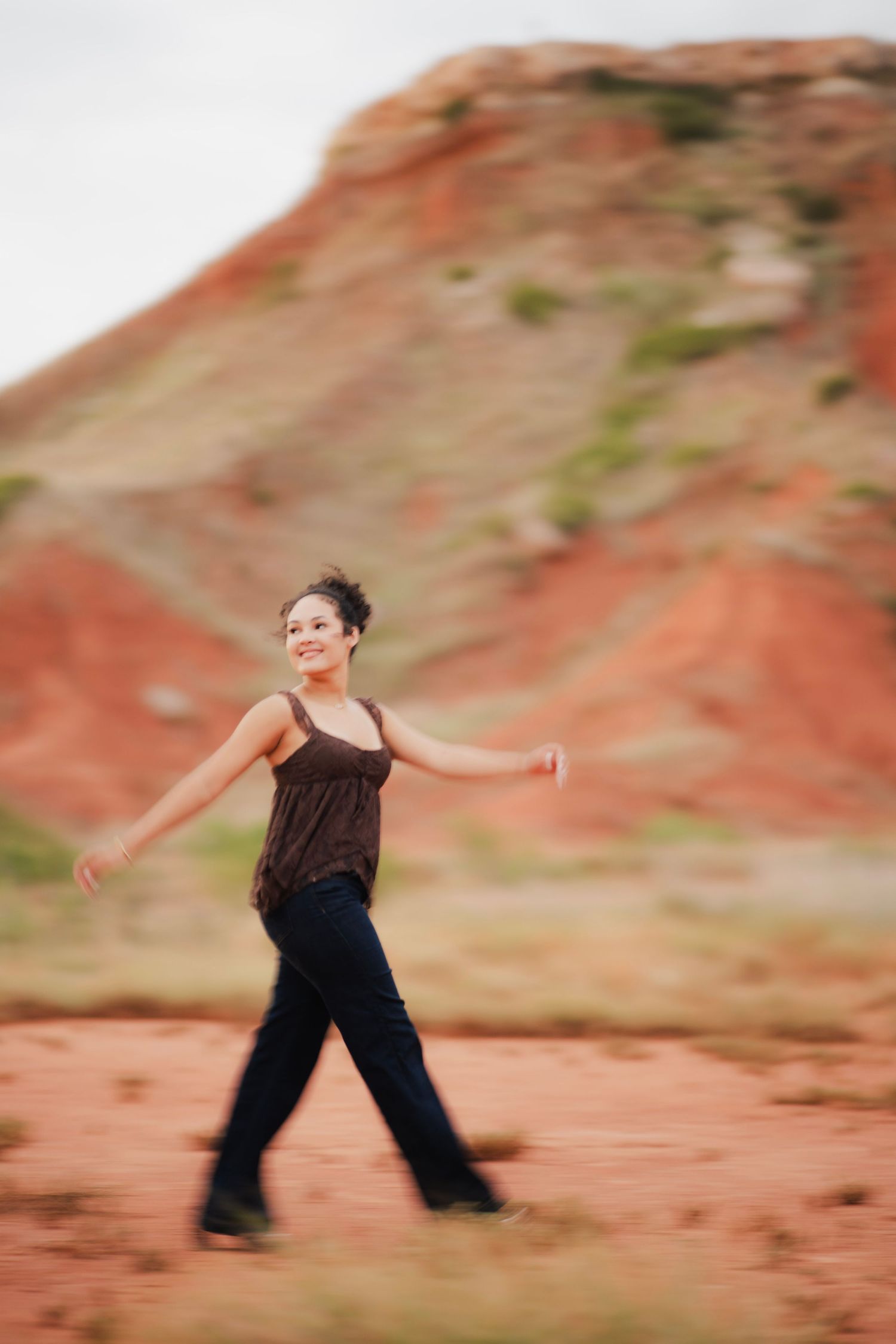 Young woman walking confidently through red rock desert landscape, wearing brown top and jeans, arms outstretched.