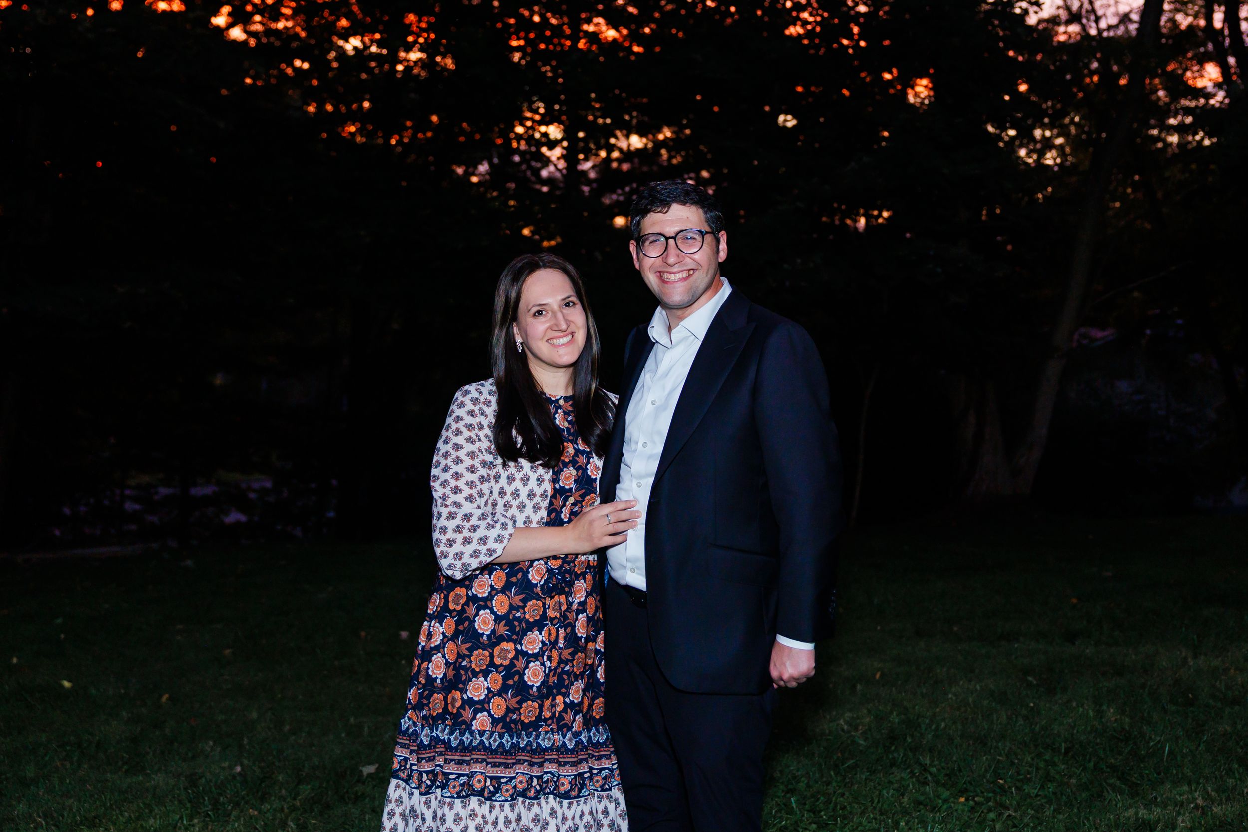 Couple smiling together outdoors at dusk, woman in floral dress and man in dark suit, trees with warm lights behind.