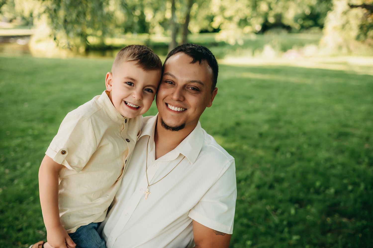 A smiling man and young boy pose together outdoors in a lush green park, wearing matching cream shirts.